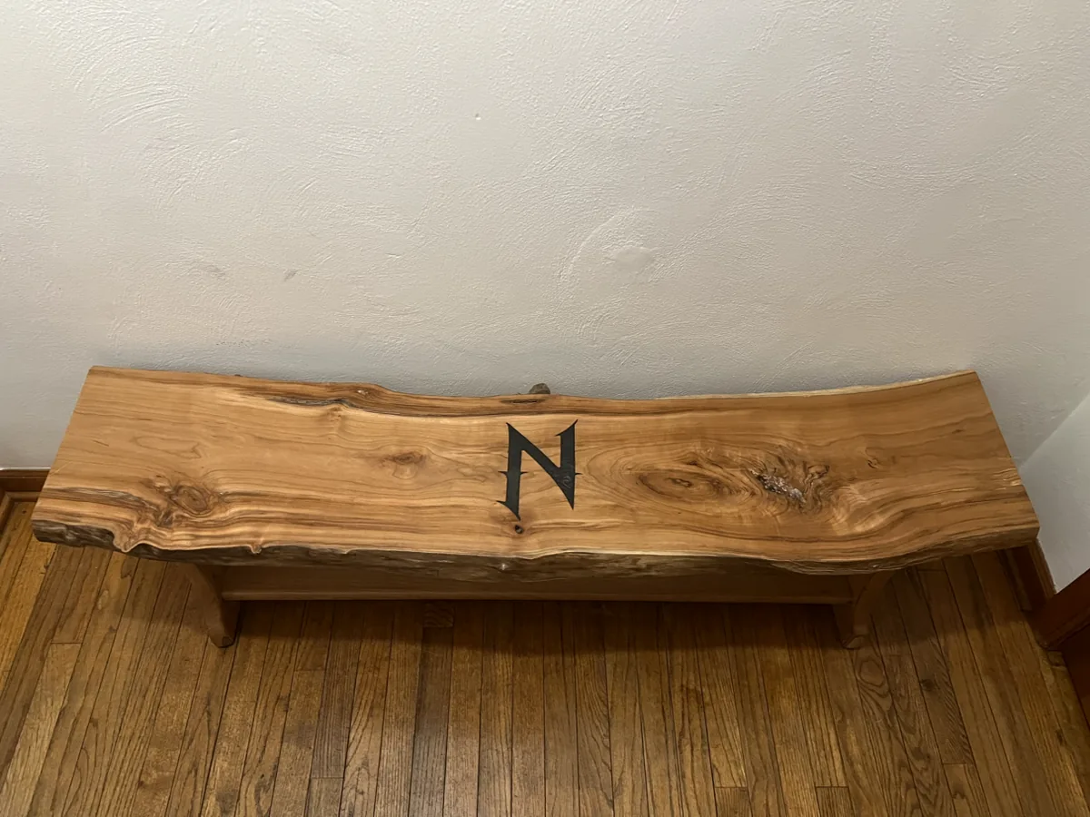 Live-edge wood bench with a dark angular design photographed against a white wall.